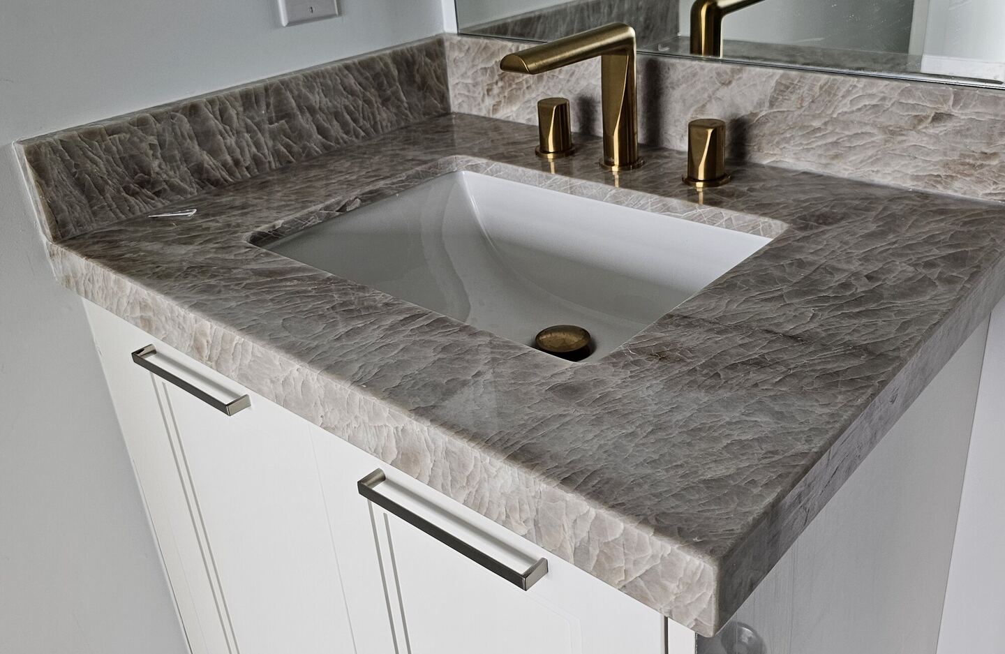 Undermount sink cut into a grey veined stone vanity top with brass faucet, installed by Pearl Countertops